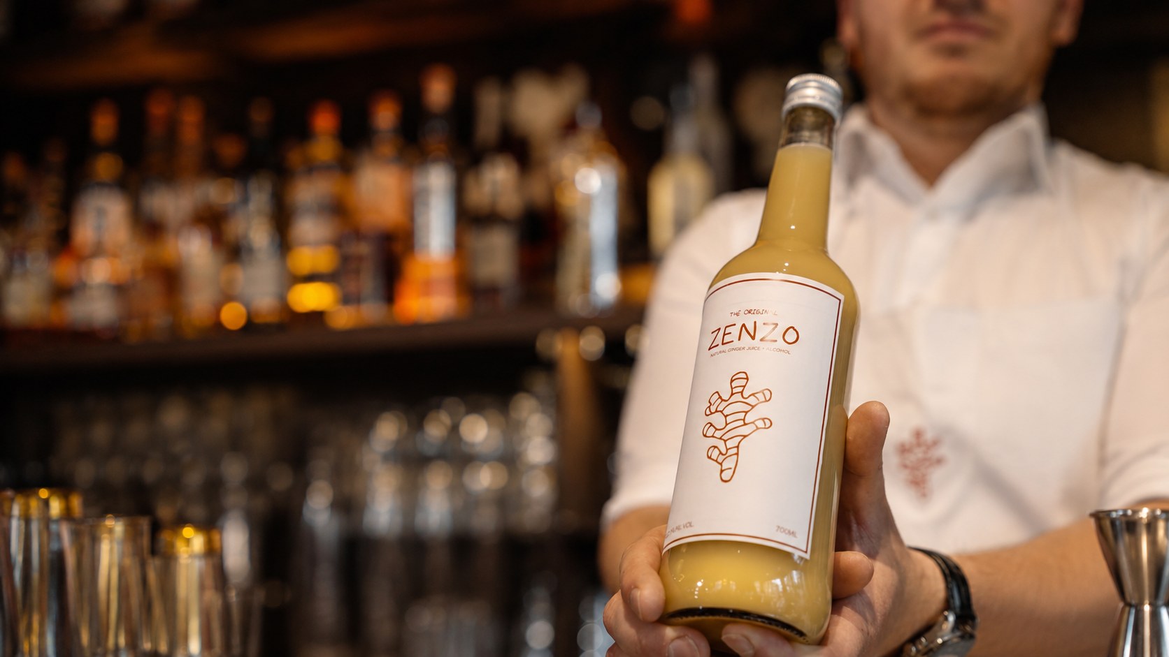 Bartender met witte polo houdt een fles Zenzo omhoog voor een verlichte bar